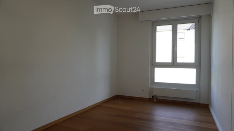 Empty room with wooden flooring, white walls, and a large window with blinds. A radiator is visible below the window.