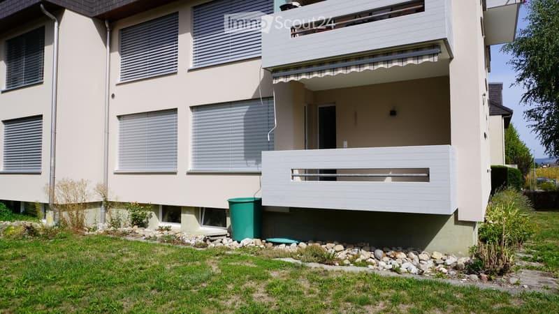 Two-story residential building with balconies, beige exterior, multiple windows with blinds, and a small garden area with rocks and greenery.