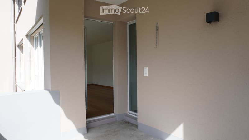 Empty room with wooden flooring, visible through an open sliding glass door from a concrete patio area.