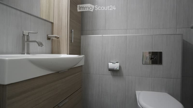 Modern bathroom with a white sink, sleek faucet, wooden cabinet, and toilet. Grey tiled walls and a mirror above the sink.