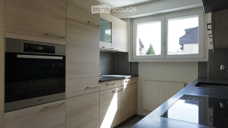 Modern kitchen with light wooden cabinets, stainless steel oven, black countertop, and a window overlooking the outside.