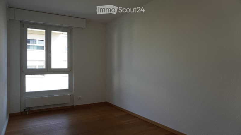 Empty room with wooden flooring, white walls, and large windows with a radiator below them.