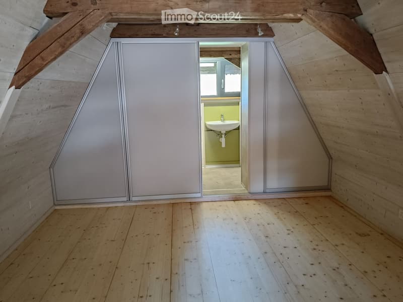 An attic space with wooden beams and flooring, featuring a sliding door that opens to a small bathroom with a sink.