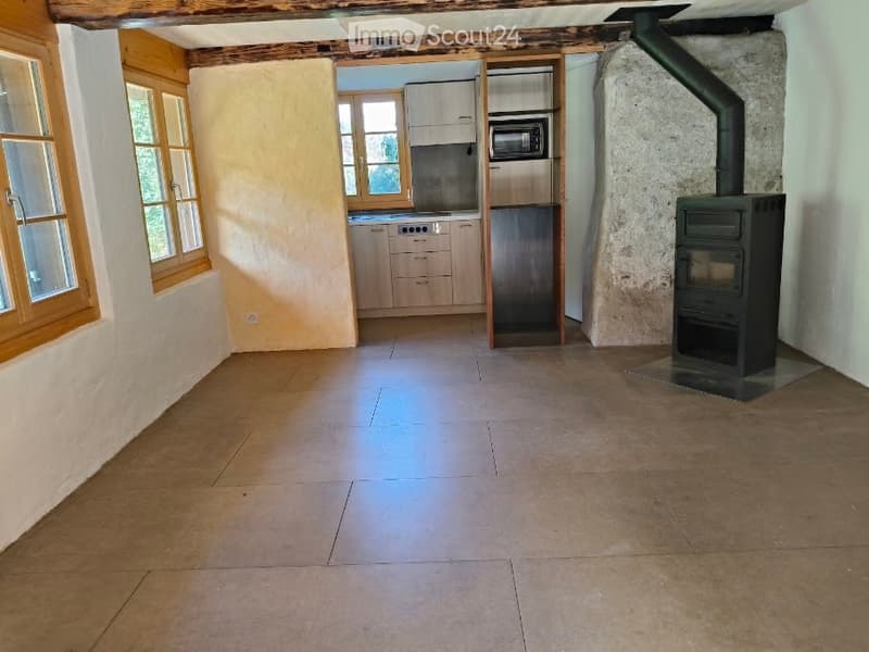 The image shows a partially furnished room with a wooden floor, wooden windows, and a wooden ceiling. There is a microwave, refrigerator, and cabinets in the background, along with a wooden stove on the right side.