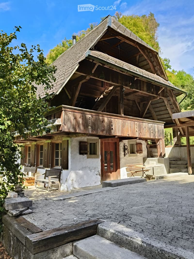 A rustic wooden house with a steep, multi-pitched roof, white plaster walls, and wooden shutters. The house features a covered porch with wooden benches and tables, surrounded by greenery and stone steps.