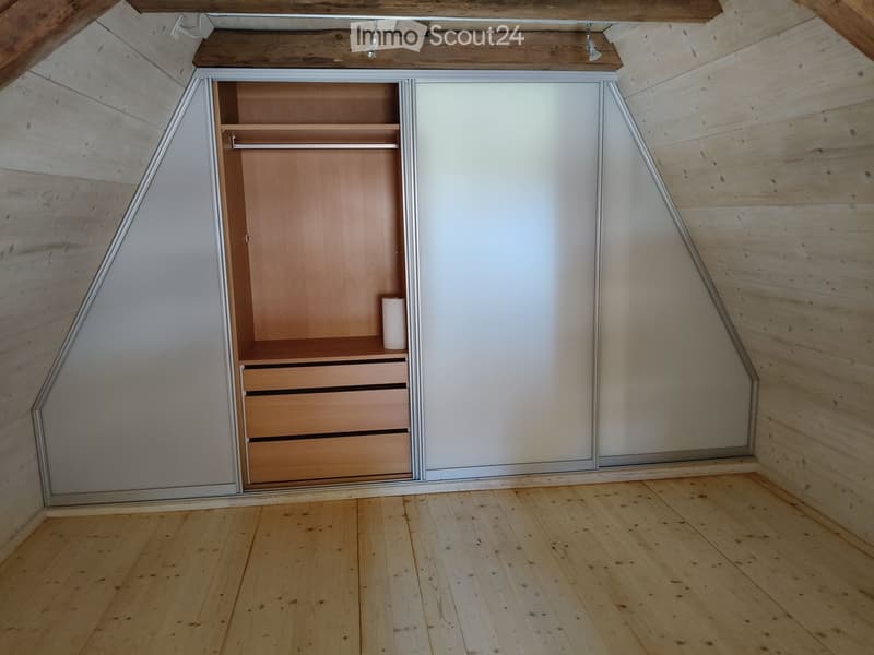 An empty attic room with wooden floors, slanted wooden ceiling, and built-in closet with shelves and drawers.