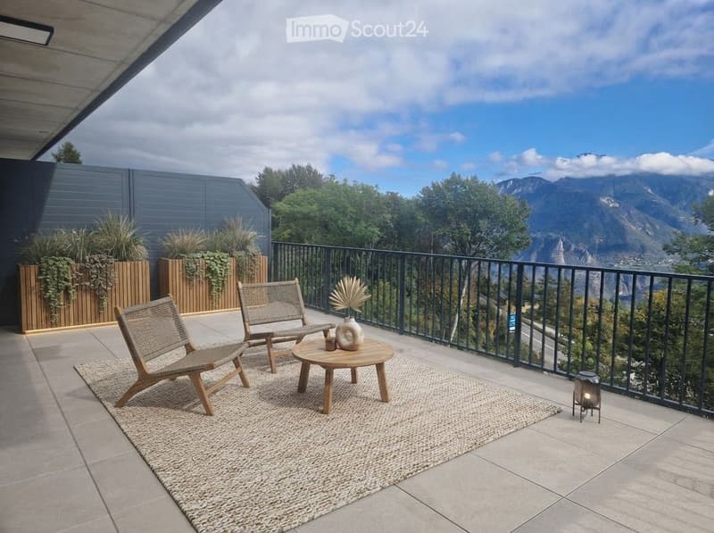 Balcony with a scenic mountain view, featuring two wooden chairs, a small round table, a decorative vase, and a woven rug.
