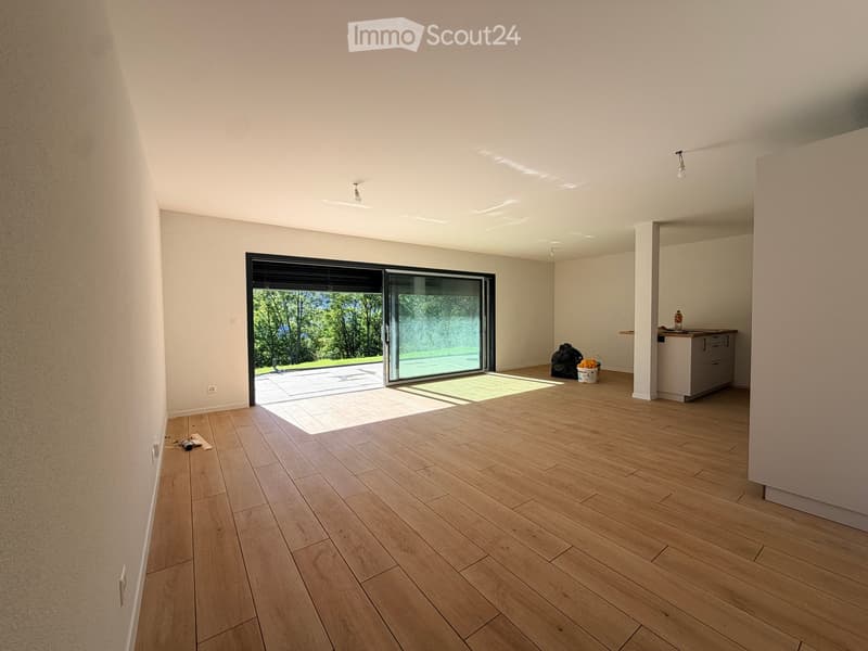 Empty room with wooden flooring, white walls, and a large sliding glass door leading to an outdoor area. There is a small kitchen area visible in the background.