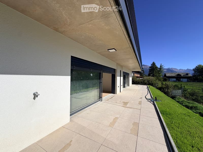 Modern outdoor patio with tiled flooring, glass sliding doors leading to a building, and a well-maintained lawn with a view of mountains in the background.