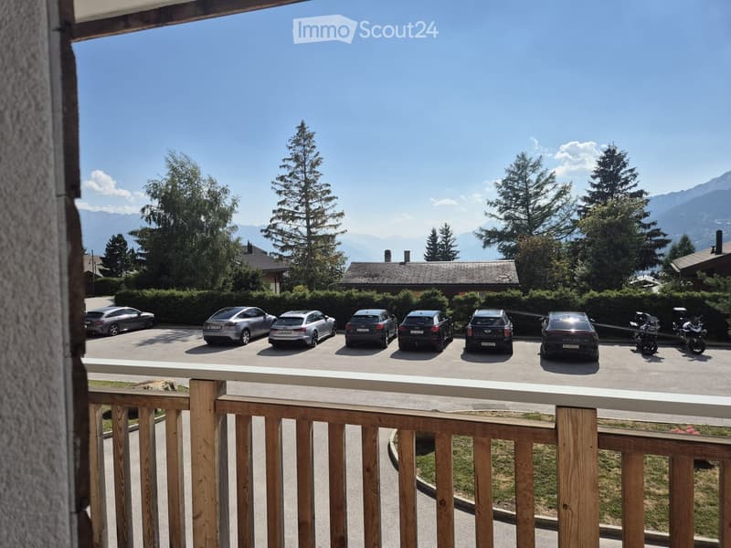 Balcony overlooking a parking area with several cars and motorcycles, surrounded by trees and mountains in the background.