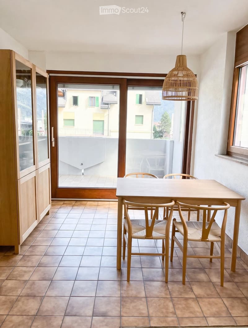 Dining area with a wooden table, four chairs, and a wooden cabinet with glass doors, overlooking a balcony.
