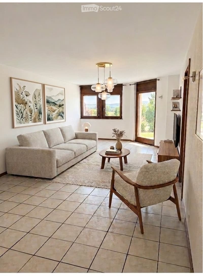 Bright living room with large windows offering a scenic view, featuring a beige sofa, wooden coffee table, armchair, table lamp, decorative vase, and artwork on the walls.