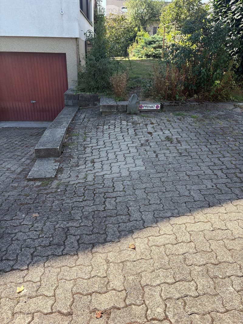 A cracked stone driveway leading to a house with a red garage door, surrounded by overgrown vegetation and a "PRIVAT" sign.