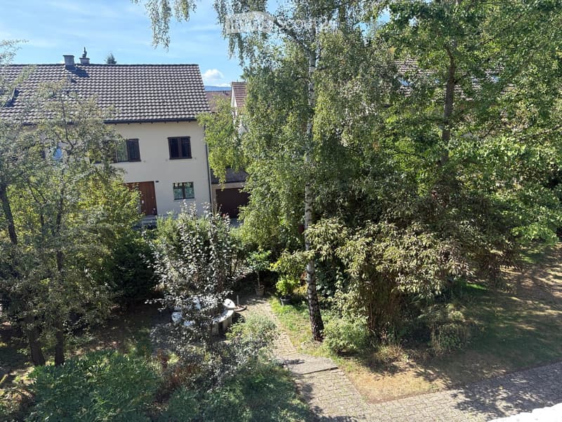 Two-story house with a tiled roof, surrounded by a lush garden with trees, shrubs, and a paved pathway.