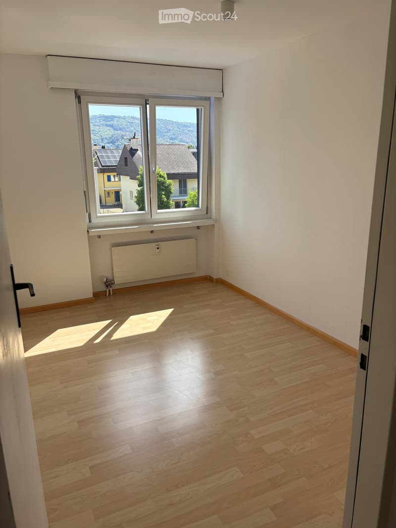 Empty room with wooden flooring, white walls, and large windows offering a view of the surrounding area.