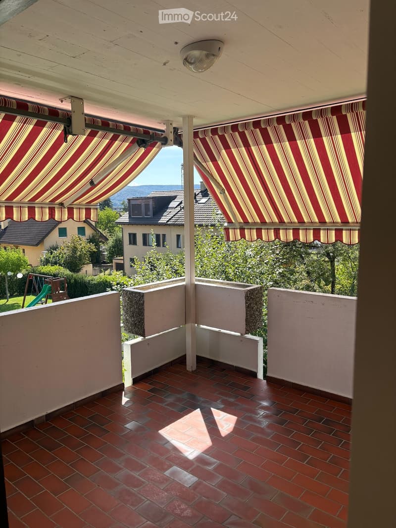Balcony with red and white striped awnings, red tiled floor, white railings, and a view of surrounding houses and greenery.