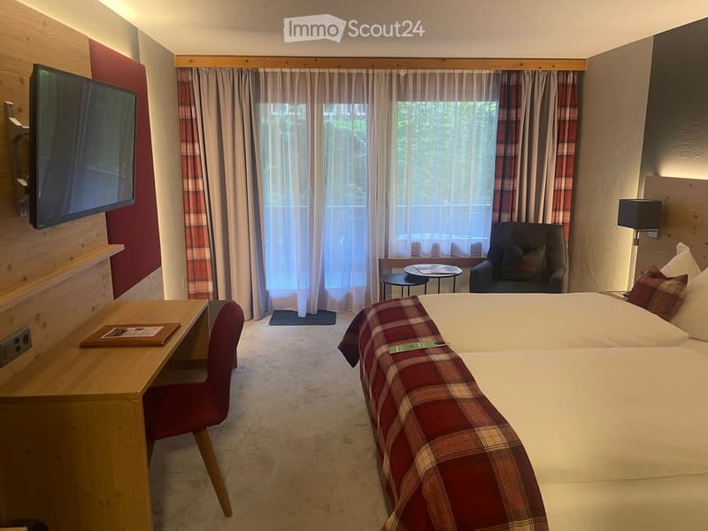 Cozy bedroom with a large bed, wooden desk, red chair, small round table, armchair, and a TV mounted on the wall. Large windows with curtains provide natural light.