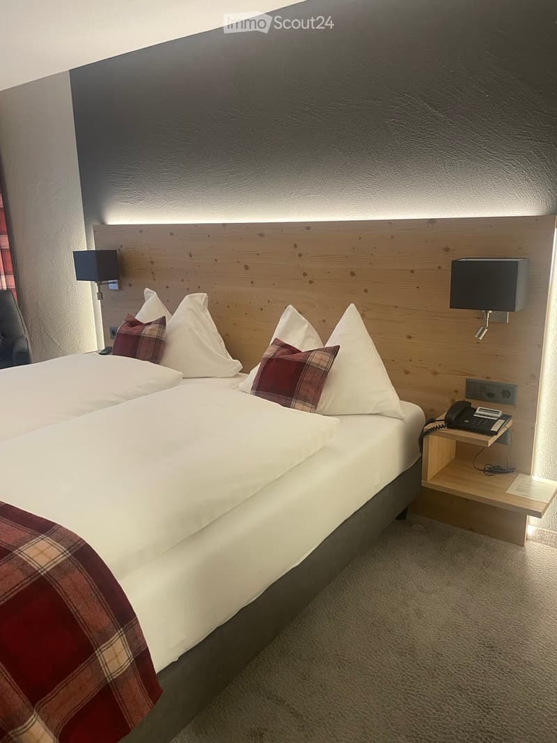A neatly made double bed with white linens, red plaid blanket, and decorative pillows, accompanied by a wooden nightstand with a telephone and lamp.