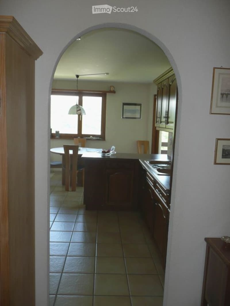 A modern kitchen with a central island, stove, and dining area. The space features wooden cabinets, a tiled floor, and large windows allowing natural light to fill the room.