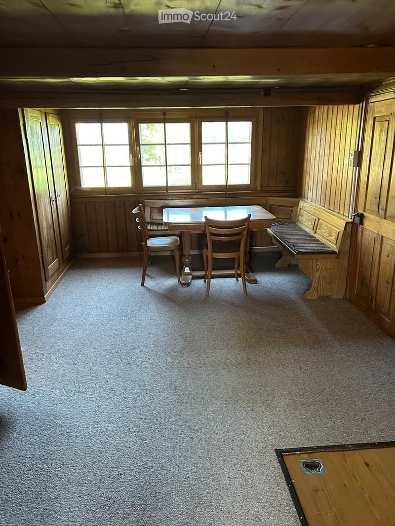 Cozy wooden-paneled room with a small dining table, two chairs, and a built-in bench along one wall. The room has large windows and carpeted flooring.