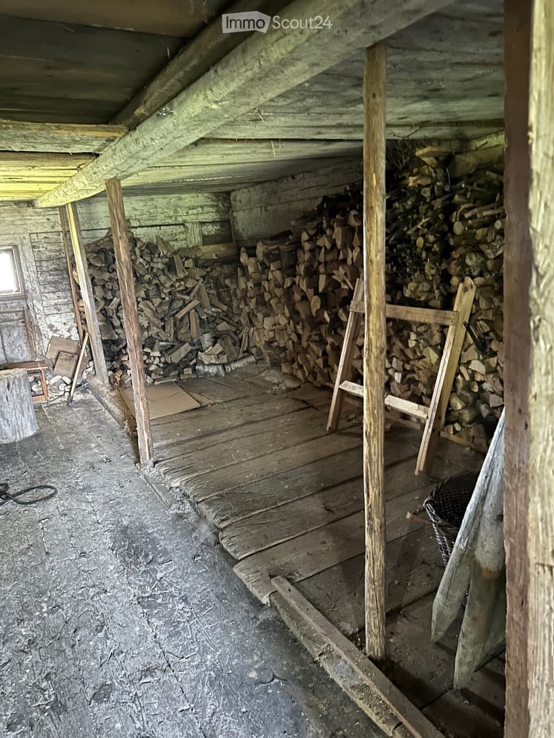 An attic or storage space with wooden beams, a concrete floor, and stacks of firewood along the walls. The space is unfinished with exposed wooden beams and a small window on one side.