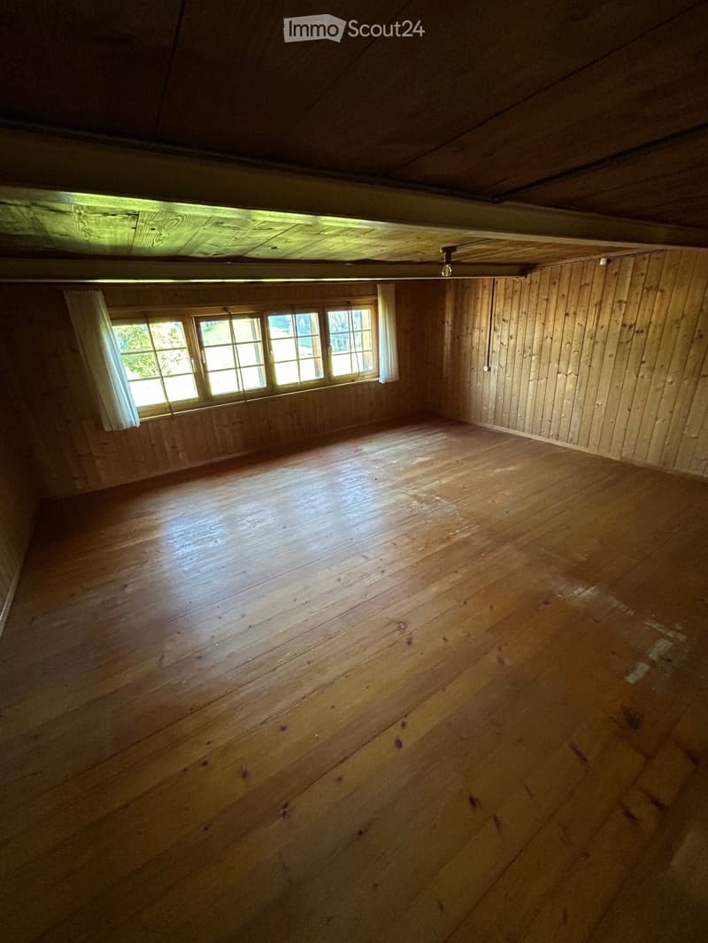 Empty wooden room with exposed wooden ceiling beams, wooden walls, and a large window with white curtains. The floor is made of wide wooden planks.