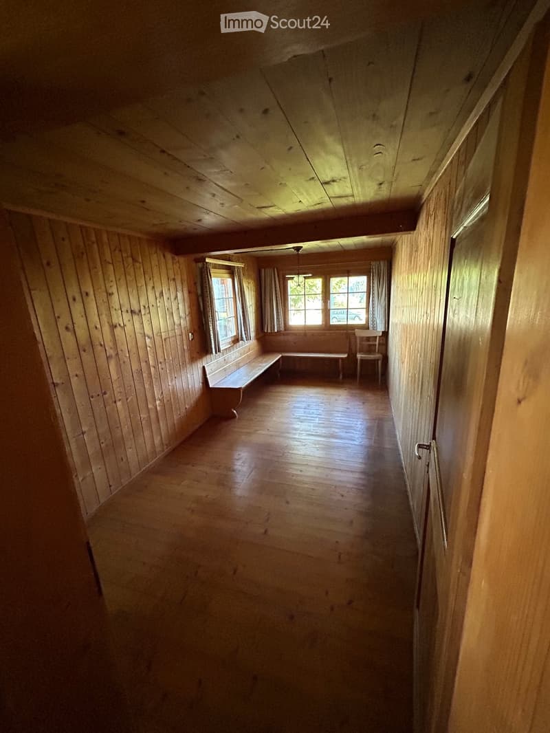 Empty wooden hallway with wooden walls, floor, and ceiling. Features a bench and a chair near a window with curtains.
