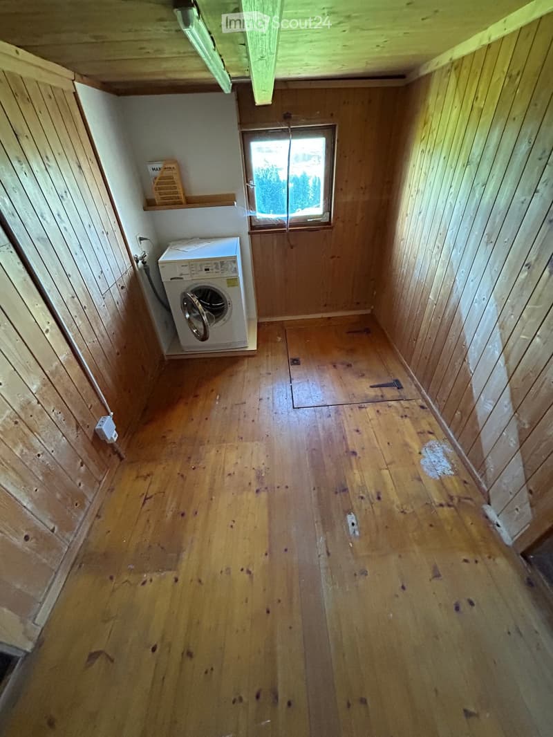 Empty wooden-floored room with a washing machine, wooden walls, and a small window showing greenery outside.