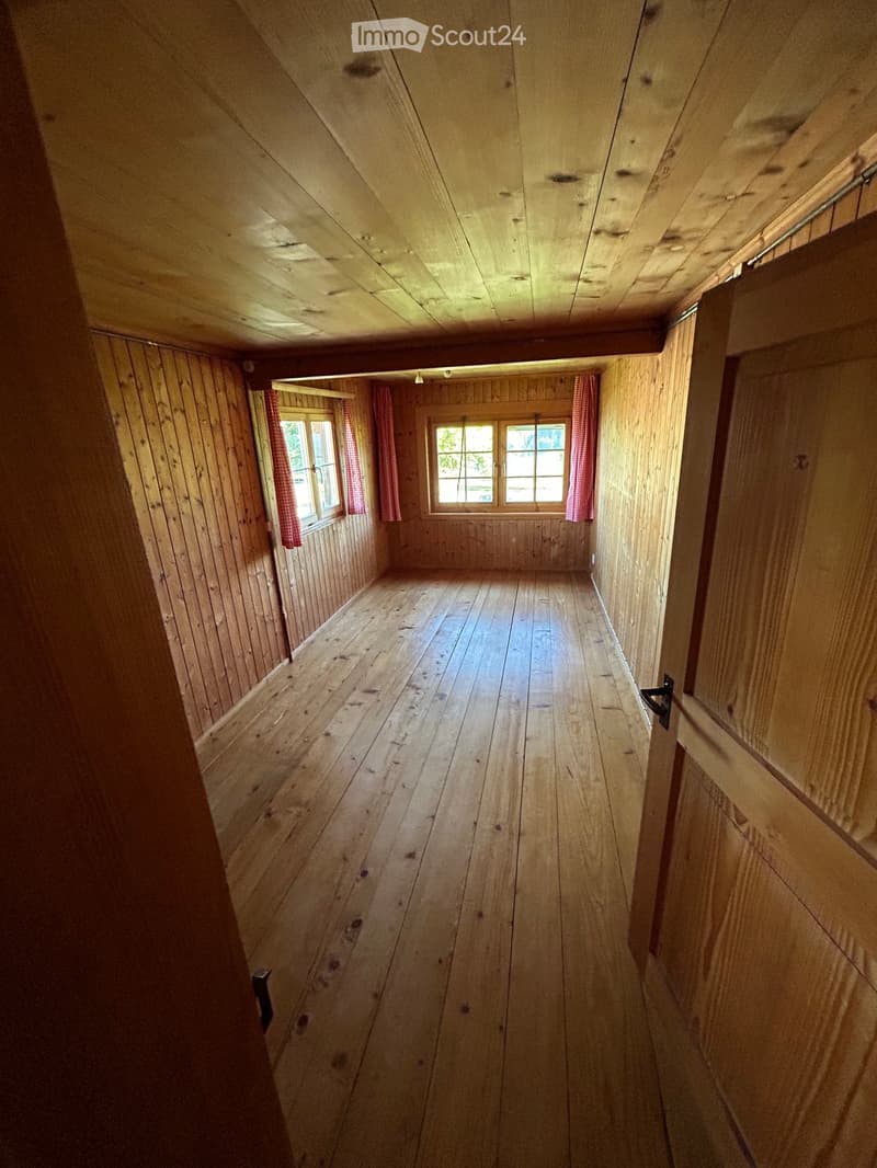 Empty wooden hallway with exposed wooden beams, wooden floor, and two windows with red checkered curtains.