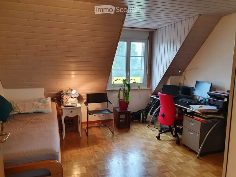 Cozy attic room with wooden walls and slanted ceilings, featuring a bed, desk with office chair, laptop, printer, and a small plant on a side table.