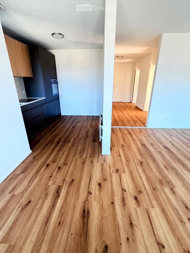Empty hallway with light blue walls and polished wooden flooring, leading to a kitchen area with dark cabinets and stainless steel appliances.