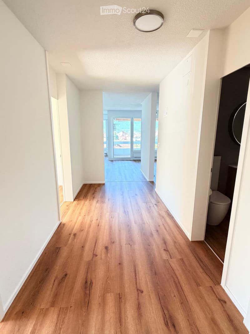 Bright hallway with wooden flooring, white walls, and recessed lighting. The hallway leads to an open area with large glass doors and a view of the outside.