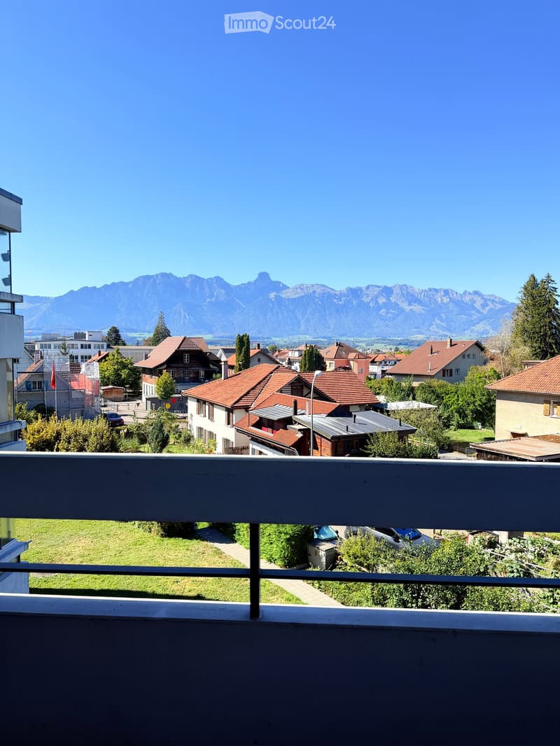 Balcony overlooking a residential area with houses, trees, and distant mountains under a clear blue sky.
