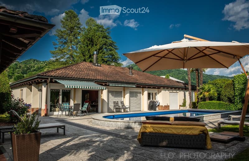Outdoor area with a swimming pool, patio furniture, umbrella, and a view of a house with a tiled roof and greenery.