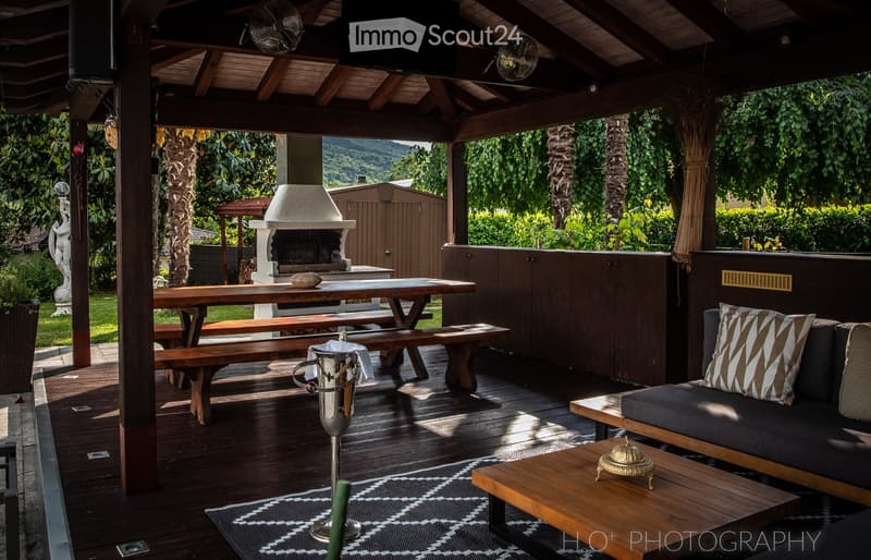 Patio with wooden flooring, outdoor seating, central fireplace, dining table, couch with cushions, metal stands, garden, hedges, green foliage, mountains in the background