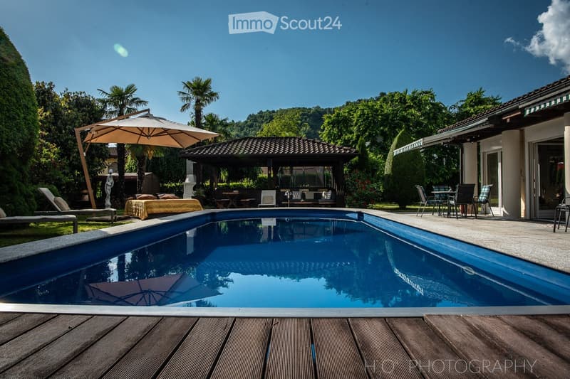 Swimming pool with wooden decking, outdoor seating with umbrella, outdoor dining table, gazebo, lawn with lounge chairs, and tropical trees.