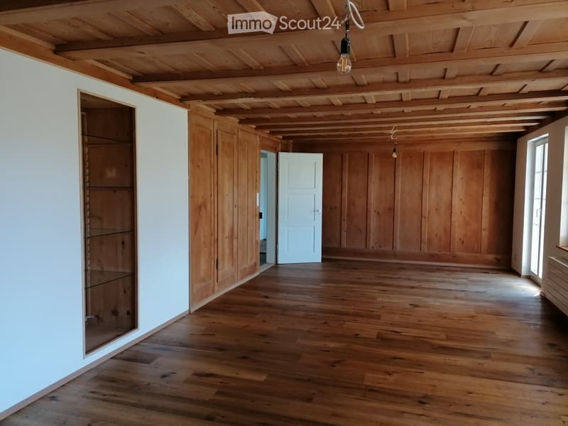 Empty room with wooden floors, white walls, exposed wooden ceiling beams, and a glass-fronted cabinet.