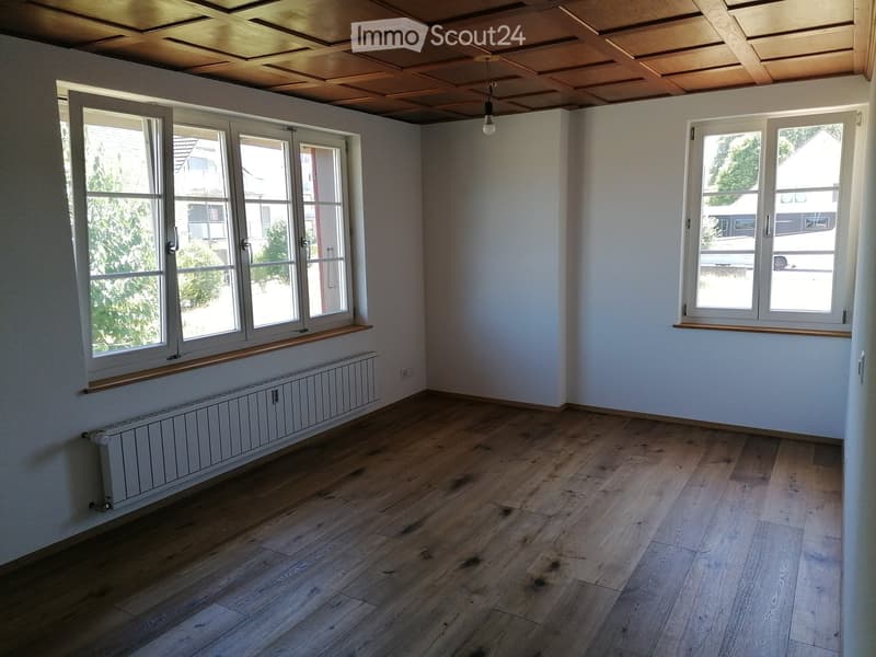 Empty room with wooden flooring, white walls, large windows, and a radiator under one window.