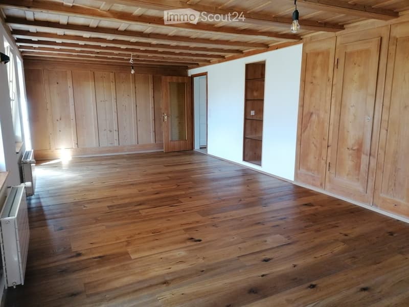 Empty room with wooden floors, exposed wooden beams on the ceiling, and large wooden doors. Natural light streams in from the left side.