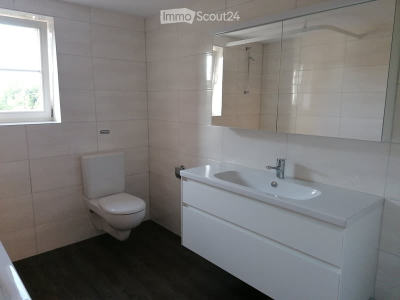 Modern bathroom with white tiles, a toilet, a sink with a modern faucet, and a large mirror above the sink. The room has a window on the left side, allowing natural light to enter.