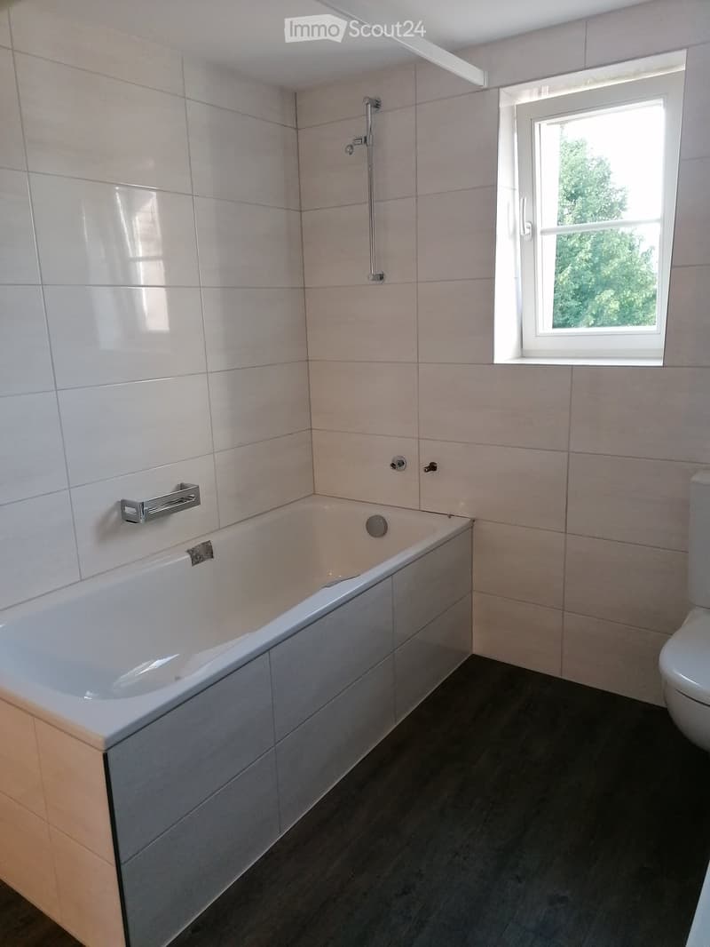 Modern bathroom with a white bathtub, toilet, shower head, and a window letting in natural light.
