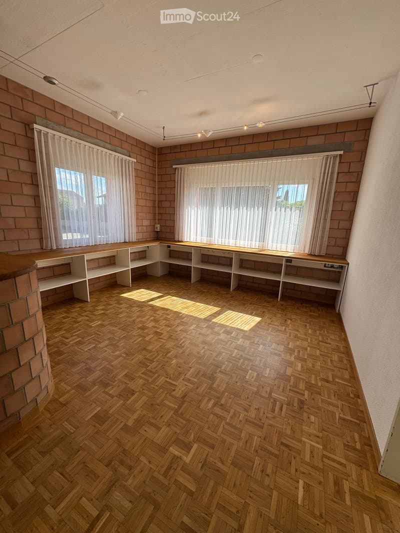 Empty room with wooden herringbone flooring, exposed brick walls, built-in shelving units, and large windows with sheer curtains.