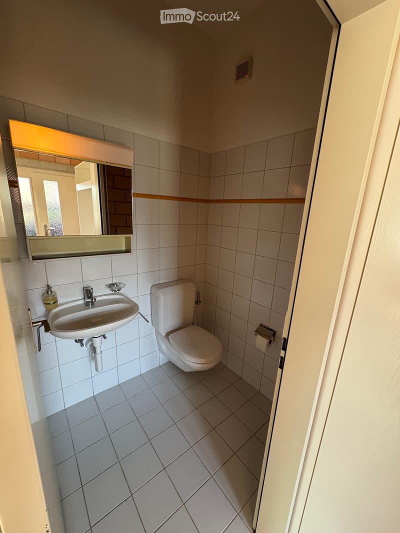 Small bathroom with white tiled walls and floor, featuring a sink, toilet, and mirror with a built-in shelf.