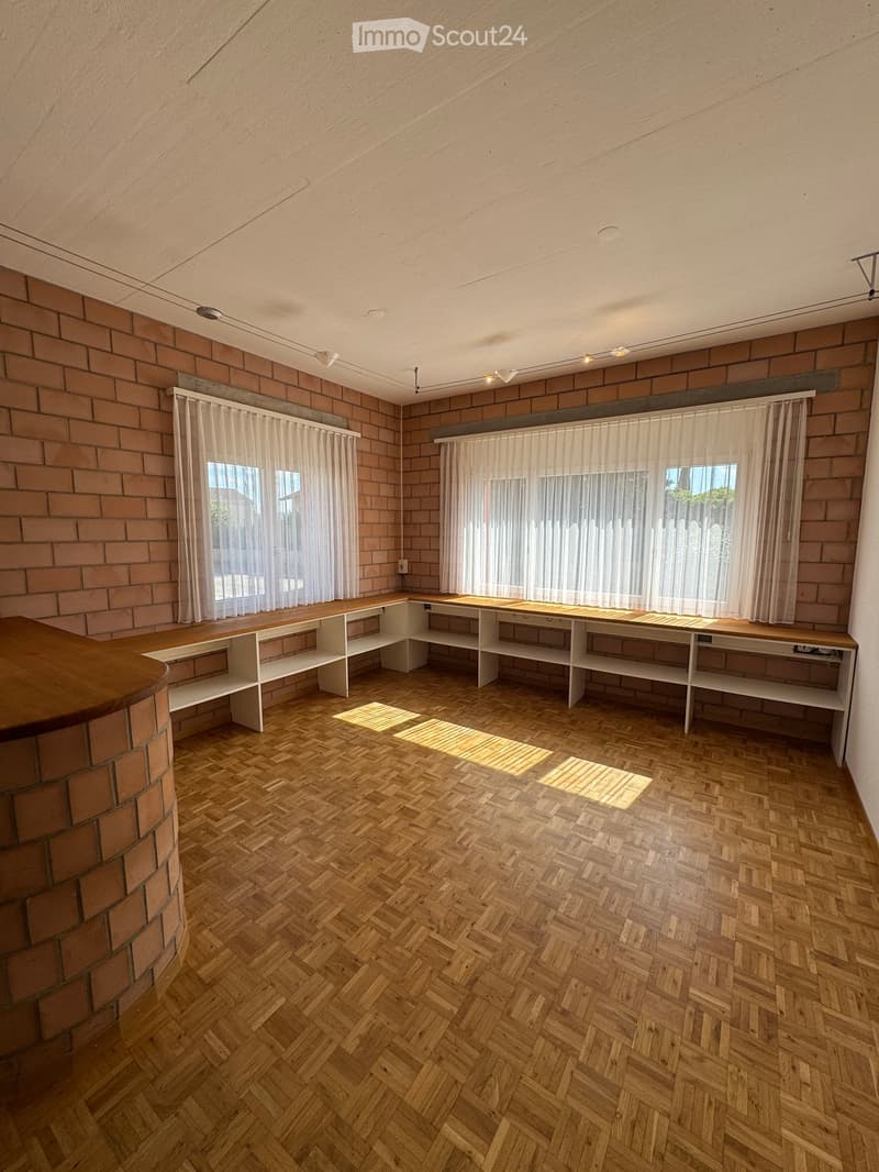 Empty room with exposed brick walls, wooden herringbone flooring, built-in shelving units, and large windows with sheer curtains.