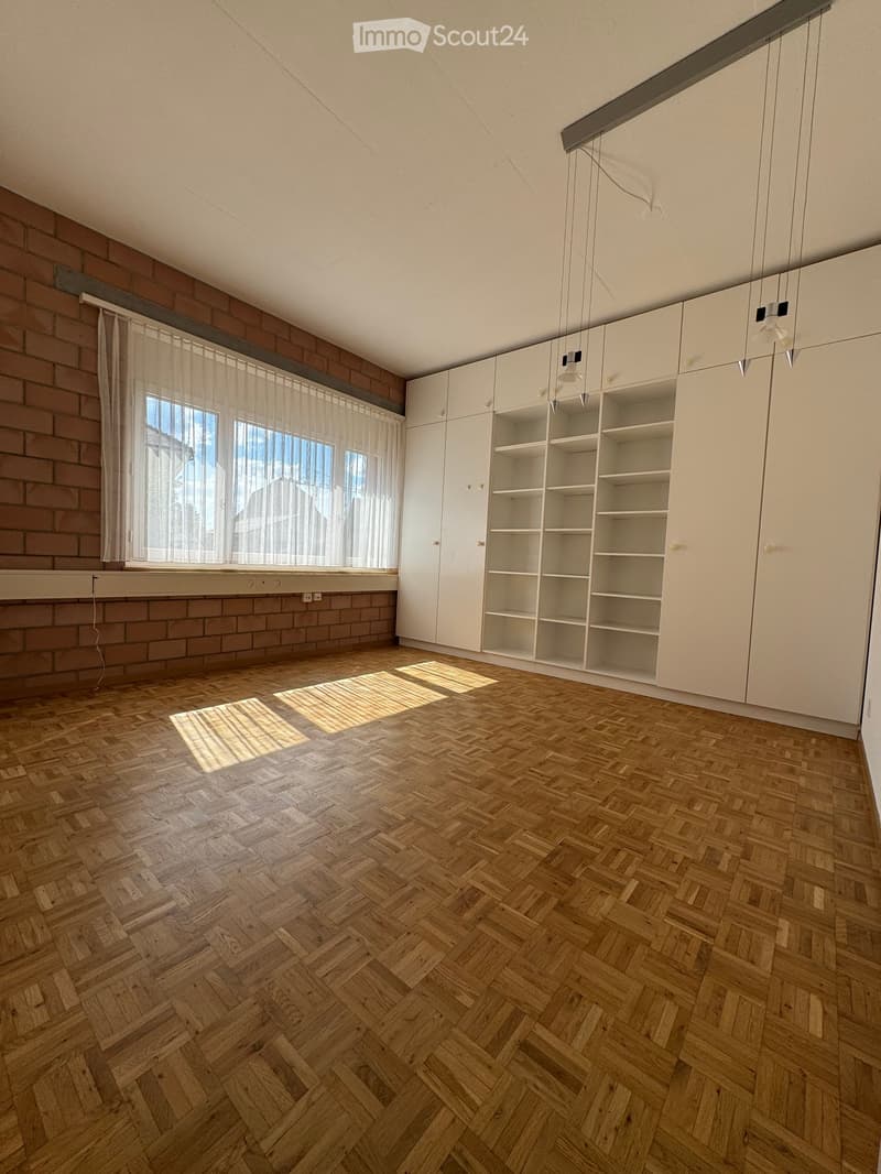 Empty room with wooden herringbone flooring, white built-in cabinets, large windows with sheer curtains, and exposed brick wall.