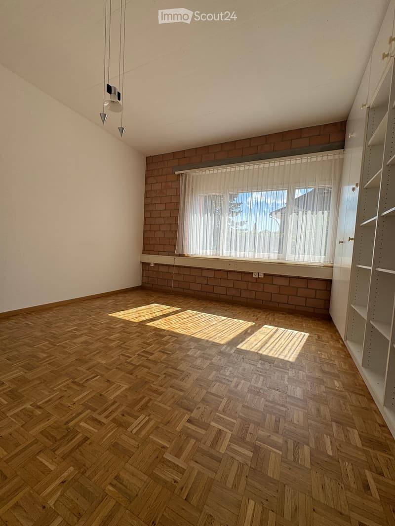 Empty room with wooden herringbone flooring, white walls, and a large window with sheer curtains. The room features exposed brickwork along one wall and built-in shelving units on the right side.