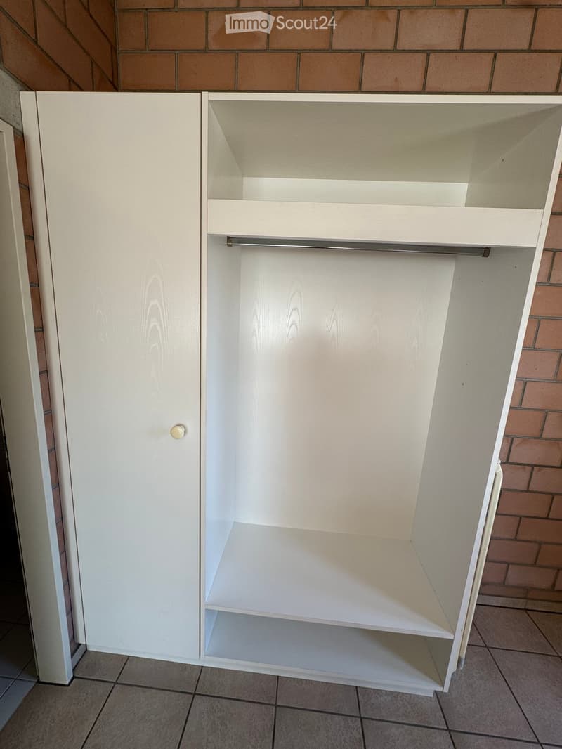 Empty room with a white built-in closet featuring shelves and a hanging rod, against a brick wall with tiled flooring.