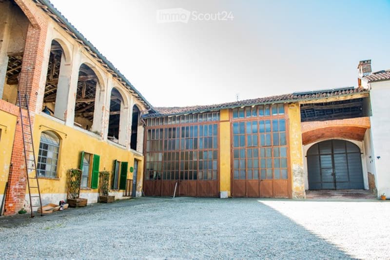 An old rustic building with arched windows and large wooden doors, featuring a gravel courtyard and some potted plants near the entrance.
