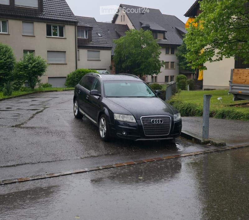 A wet residential street with a parked Audi SUV in front of a multi-story house with multiple windows and a sloped roof.