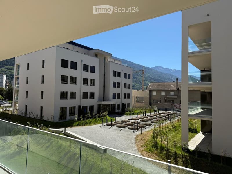 Modern multi-story building with white facade and numerous windows, surrounded by landscaped area with seating and greenery, with mountains in the background.